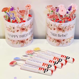 Op de afbeelding: Twee cilindrische geschenkdozen versierd met bloemmotieven en de zinnen "HAPPY BIRTHDAY" en "Mit liebe". Verschillende chocoladerepen met bloemtoppers zijn ervoor geplaatst.