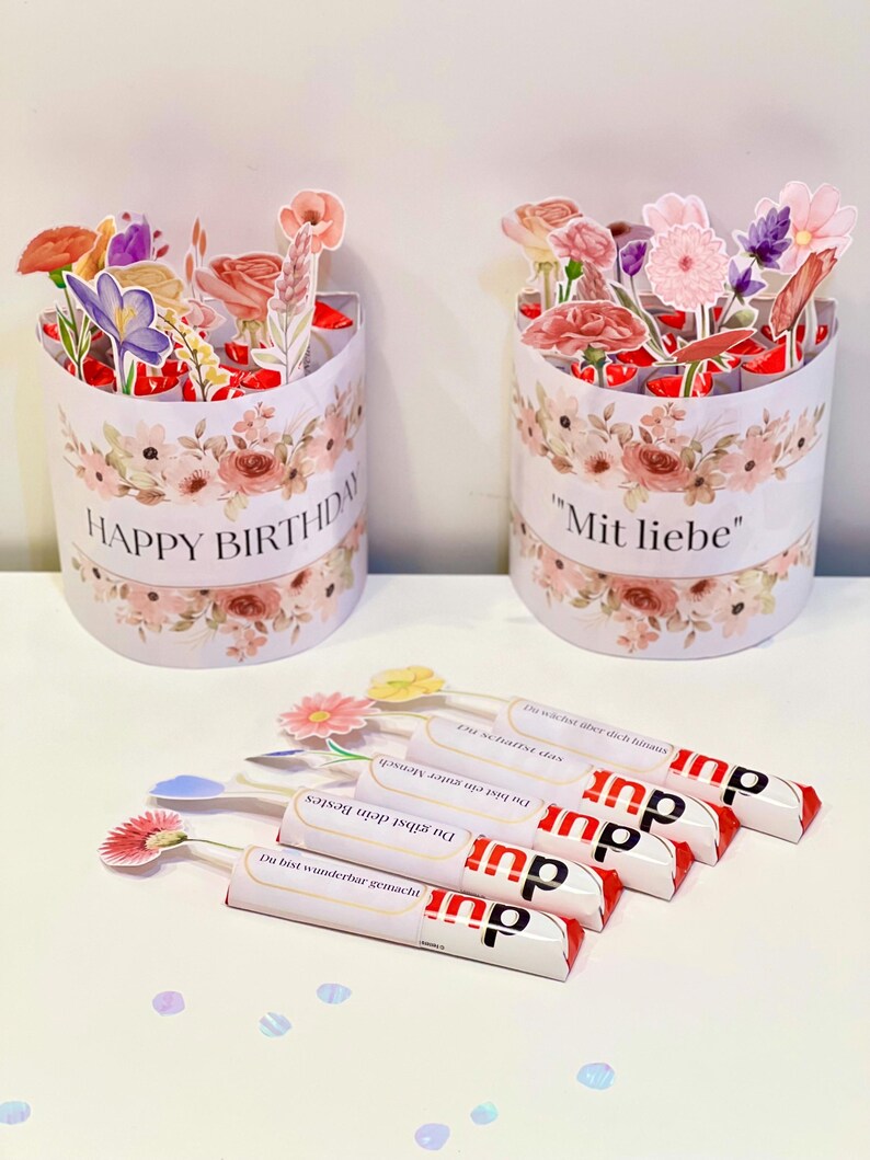 Op de afbeelding: Twee cilindrische geschenkdozen versierd met bloemmotieven en de zinnen "HAPPY BIRTHDAY" en "Mit liebe". Verschillende chocoladerepen met bloemtoppers zijn ervoor geplaatst.