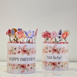 Op de afbeelding: Twee cilindrische geschenkdozen met bloemmotieven en papieren bloemstukken. E&eacute;n doos leest "HAPPY BIRTHDAY" in zwarte tekst. De andere doos leest "Mit liebe" in zwarte tekst. De dozen hebben een witte achtergrond met roze en perzik bloemen.