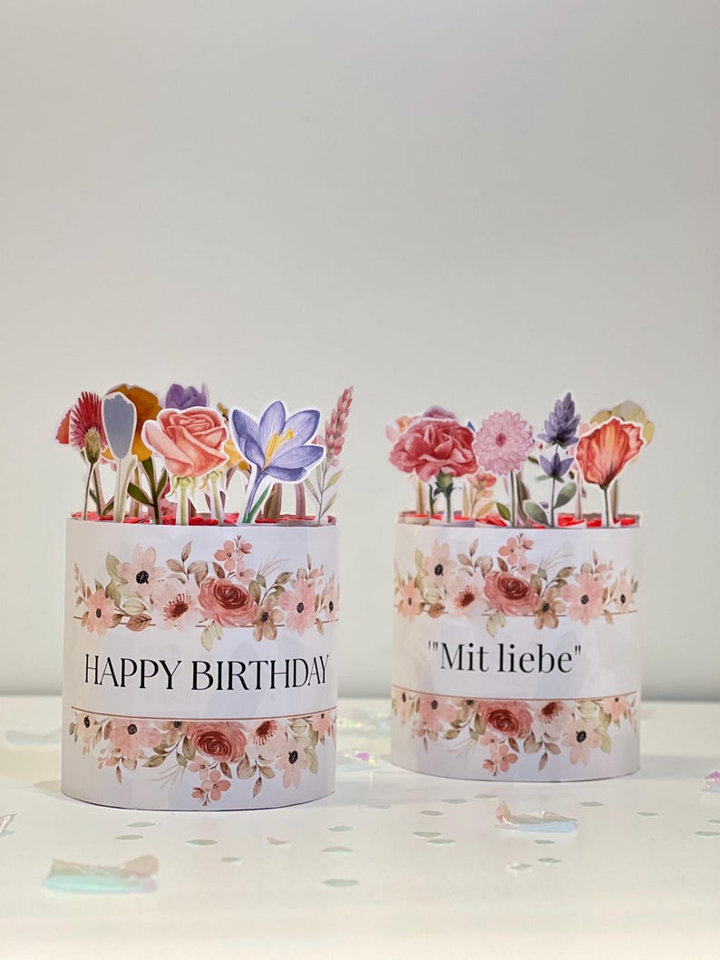 Op de afbeelding: Twee cilindrische geschenkdozen met bloemmotieven en papieren bloemstukken. E&eacute;n doos leest "HAPPY BIRTHDAY" in zwarte tekst. De andere doos leest "Mit liebe" in zwarte tekst. De dozen hebben een witte achtergrond met roze en perzik bloemen.