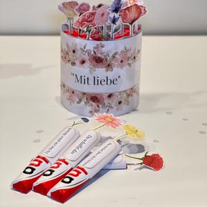 Op de afbeelding: Een bloemengeschenkset. Het bevat een cilindrische container met de tekst "Mit liebe" en papieren bloemen. Er zijn ook drie chocoladerepen met Duitse tekst aanwezig, samen met extra papieren bloemen.