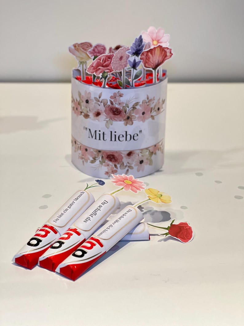 Op de afbeelding: Een bloemengeschenkset. Het bevat een cilindrische container met de tekst "Mit liebe" en papieren bloemen. Er zijn ook drie chocoladerepen met Duitse tekst aanwezig, samen met extra papieren bloemen.