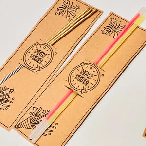 May include: Brown New Year's party favours with a clock design and the words "NEW YEAR". One favour has gold sticks, and the other has pink and yellow glow sticks. The favours have a dotted border and starburst designs.