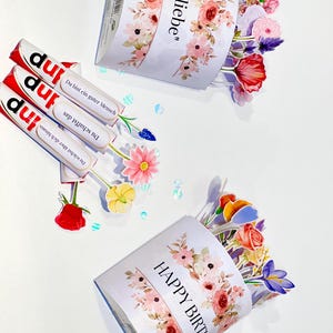 Op de afbeelding: Een witte geschenkdoos met bloemmotieven en de tekst "Happy Birthday" en "Mit liebe". De doos is gevuld met kleurrijke papieren bloemen. Er zijn ook drie chocoladerepen met Duitse tekst.