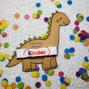 May include: A brown dinosaur-shaped invitation with the text "Einladung zu meinem Geburtstag" and a Kinder chocolate bar. Colorful confetti in shades of blue, orange, yellow, and purple surrounds the dinosaur on a white textured surface.
