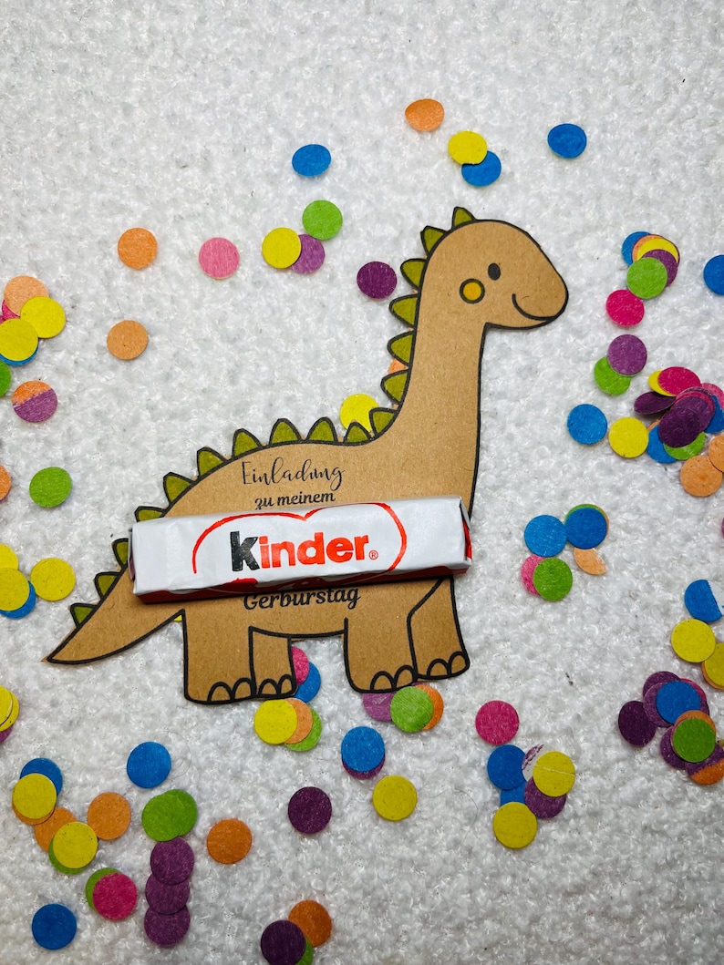 May include: A brown dinosaur-shaped invitation with the text "Einladung zu meinem Geburtstag" and a Kinder chocolate bar. Colorful confetti in shades of blue, orange, yellow, and purple surrounds the dinosaur on a white textured surface.