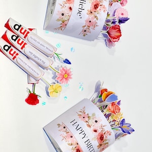 Op de afbeelding: Een witte geschenkdoos met bloemmotieven en de tekst "Happy Birthday" en "Mit liebe". De doos is gevuld met kleurrijke papieren bloemen. Er zijn ook drie chocoladerepen met Duitse tekst aanwezig.