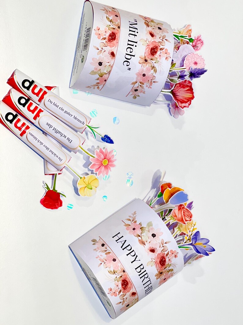 Op de afbeelding: Een witte geschenkdoos met bloemmotieven en de tekst "Happy Birthday" en "Mit liebe". De doos is gevuld met kleurrijke papieren bloemen. Er zijn ook drie chocoladerepen met Duitse tekst aanwezig.