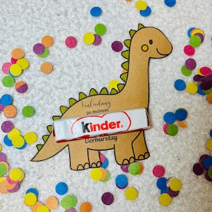 May include: A brown dinosaur-shaped invitation with the text "Einladung zu meinem Kinder Geburtstag" and a Kinder chocolate bar. Colorful confetti in various colors surrounds the invitation on a white surface.