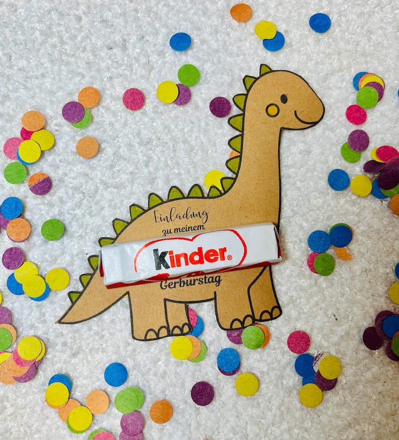 May include: A brown dinosaur-shaped invitation with the text "Einladung zu meinem Kinder Geburtstag" and a Kinder chocolate bar. Colorful confetti in various colors surrounds the invitation on a white surface.