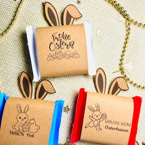 Puede incluir: Barras de chocolate con temática de Pascua envueltas en papel marrón con bordes coloridos. Los envoltorios presentan ilustraciones de conejitos y texto en alemán como "Frohe Ostern" y "Gruss vom Osterhasen". Orejas de conejo decorativas y cuentas doradas completan la escena festiva.