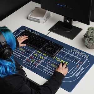 May include: A blue gaming mouse pad with a craps table design, featuring white lines and text. The pad includes the words "COME", "PASS LINE", and "FIELD". A black keyboard and mouse are on the pad.