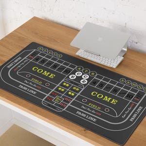 May include: A black craps table mat with white and yellow text, including "COME", "FIELD", and "PASS LINE". The mat features dice combinations and numbers from 4 to 12. A laptop and keyboard rest on a wooden surface above the mat.