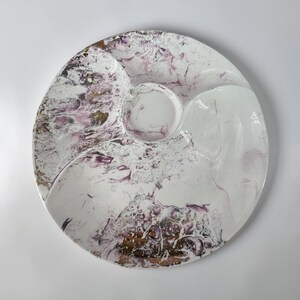 Puede incluir: Una bandeja de servicio redonda de cerámica con un diseño dividido y un compartimento central circular. La superficie muestra un patrón jaspeado en tonos blanco, morado intenso y oro metálico, creando una estética fluida y abstracta en el plato.