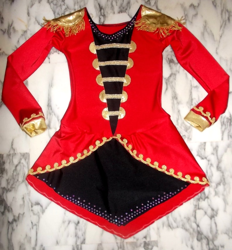 May include: Red and black leotard with gold trim and embellishments. The leotard has a long sleeve design and a skirt with a scalloped edge.