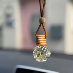 Può includere: Un piccolo flacone in vetro trasparente con un design a coste e spirali, sormontato da un tappo in legno chiaro. Il flacone è appeso a un cordino marrone con una piccola perla di legno rotonda, pensato per essere appeso in un veicolo o spazio.