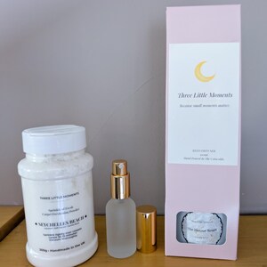 Può includere: Una collezione di prodotti per la cura di sé, tra cui un barattolo bianco di polvere rinfrescante per tappeti, un flacone spray in vetro smerigliato con tappo dorato e una scatola rosa con un logo a forma di luna e il testo "Three Little Moments."