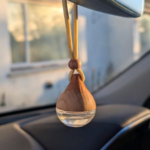 May include: A car air freshener, teardrop-shaped, with a wooden top and a clear glass base. It hangs from a yellow cord attached to the rearview mirror. The interior of the car is visible, with a blurred background of a building.