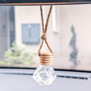 Può includere: Un profumatore per auto in vetro trasparente e sfaccettato con un tappo in legno e un cordino intrecciato marrone e oro per appenderlo. Il profumatore è appeso allo specchietto retrovisore di un'auto.