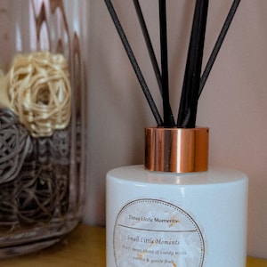 May include: A white reed diffuser with a copper-coloured collar and black reeds. The label reads "Three Little Moments" and describes a vanilla and gentle fruit scent. A glass vase with decorative spheres is in the background.
