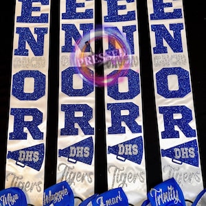 May include: White satin sashes with blue glitter lettering that reads "SENIOR" and "DHS Tigers". Each sash has a blue bow with a name embroidered on it. The sashes are for a graduation or senior event.