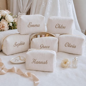 Pode incluir: Uma coleção de seis nécessaires de tecido canelado em tom branco sujo com fechos dourados. Cada nécessaire apresenta um nome bordado em letra cursiva castanha: Emma, Chloe, Sophia, Grace, Olivia e Hannah. Uma das nécessaires está aberta.