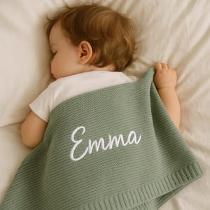 Op de afbeelding: Een slapende baby is bedekt met een saliegroene gebreide deken. De deken heeft de naam "Emma" geborduurd in witte letters. De baby draagt een witte romper en ligt op een wit laken.