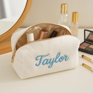 Puede incluir: Un neceser de maquillaje de pana blanca con cremallera dorada y el nombre "Taylor" bordado en azul. La bolsa está abierta, revelando varios artículos de cosmética. En el fondo se ven brochas y frascos de maquillaje.