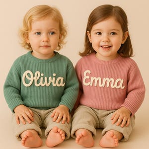 May include: Two toddlers wearing matching sweaters. One sweater is green with the name "Olivia" in white script, and the other is pink with "Emma". Both children are wearing beige pants and are sitting down.