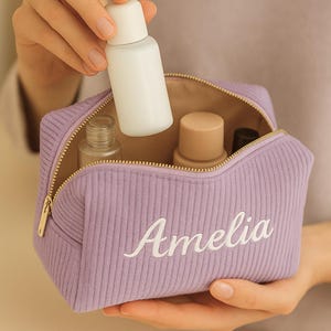 Puede incluir: Un neceser de cosméticos de pana lavanda con cremallera dorada, bordado con el nombre "Amelia" en escritura blanca. La bolsa está abierta, revelando varias botellas de cosméticos. Una mano sostiene una botella blanca sobre la bolsa.
