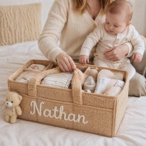 Puede incluir: Un organizador de pañales para bebé de tela sherpa texturizada en color beige con múltiples compartimentos interiores. El frente presenta el nombre Nathan bordado en letras cursivas blancas. Un pequeño oso de peluche marrón claro está sujeto al lado.