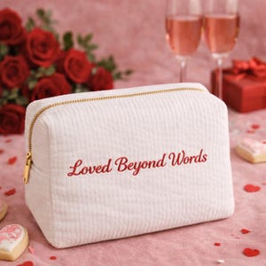 May include: A white corduroy cosmetic bag with a gold zip and the embroidered text "Loved Beyond Words" in red. The rectangular bag is on a pink surface, with red roses and champagne flutes in the background.