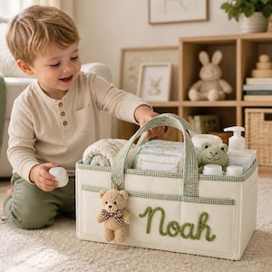 May include: A rectangular off-white fabric storage caddy with green gingham trim and handles. The front features the name Noah in green cursive embroidery and a small plush teddy bear keychain. It contains baby supplies including a frog toy.