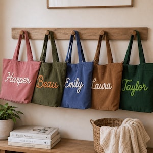 May include: Five corduroy tote bags in pink, olive green, blue, brown, and dark green hanging on a wooden rack. Each bag features a name embroidered in cursive script: Harper, Beau, Emily, Laura, and Taylor, using contrasting thread colours.