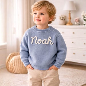 May include: A light blue long-sleeved knit jumper featuring a crew neckline and ribbed cuffs and hem. The front of the jumper displays the name Noah in white, textured, cursive-style lettering stitched onto the centre of the chest.