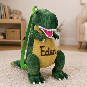 May include: A green dinosaur-shaped backpack with a yellow ribbed belly and the name "Eden" embroidered on it. The dinosaur has a wide open mouth with white teeth and a pink interior. It has green straps.