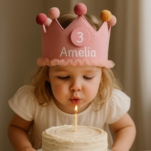 Könnte beinhalten: Ein Kind trägt eine rosa Geburtstagskrone mit dem Namen "Amelia" und der Zahl "3". Das Kind pustet eine Kerze auf einer weiß glasierten Torte aus. Die Krone hat rosa Pompons und eine Rüschenborte.