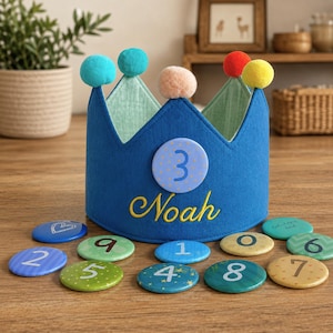 May include: A blue felt crown featuring a light blue circular patch with the number 3 and the name Noah embroidered in yellow thread. The crown is topped with five colorful pom-poms and is surrounded by several circular discs with numbers.