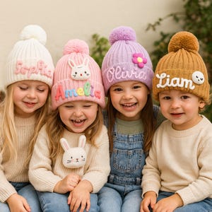 May include: Four children wearing colourful knitted beanies with pom-poms. The beanies are in white, pink, purple, and tan, each with a name embroidered on the front. The pink beanie has a bunny face. The children are smiling.