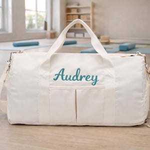 Peut inclure: Sac de sport crème avec le nom "Audrey" brodé en script turquoise. Le sac a une poignée supérieure et une bandoulière amovible. Il comporte deux poches avant et une quincaillerie en métal. Idéal pour transporter des articles de sport ou de voyage.