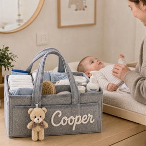 May include: A grey felt baby caddy with a gingham handle and trim, filled with baby essentials. The caddy has a teddy bear charm and the name "Cooper" embroidered on the front. Diapers, wipes, and lotion are visible.