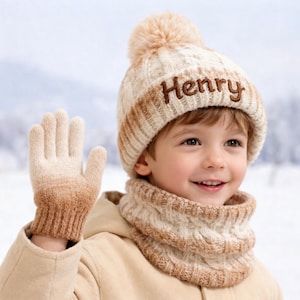 May include: A cream and brown knitted winter set featuring a hat embroidered with the name "Henry", a matching scarf, and gloves. The hat has a pom-pom. The gloves and hat have a gradient colour effect. The set is designed for cold weather.