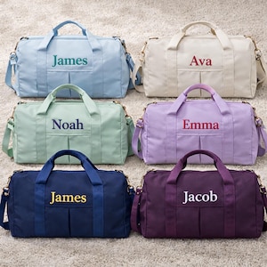 May include: Six personalised duffel bags in pastel shades: light blue, cream, mint green, lilac, navy, and deep purple. Each bag features a matching handle and shoulder strap, with a name embroidered on the front. The bags are displayed on a neutral carpet.