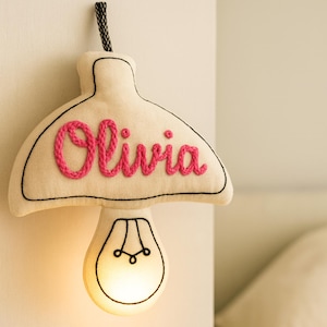 May include: A cream-colored, mushroom-shaped wall lamp with the name "Olivia" embroidered in pink. The lamp has a black outline and a lightbulb design at the bottom. A black and white cord hangs from the top.