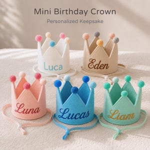 Può includere: Cinque mini corone di compleanno in feltro bianco, beige, rosa, blu e verde menta, ognuna con un nome diverso ricamato sul davanti. Ogni corona ha un pompon su ogni punta e un sottogola regolabile. Il testo in alto dice "Mini Birthday Crown".