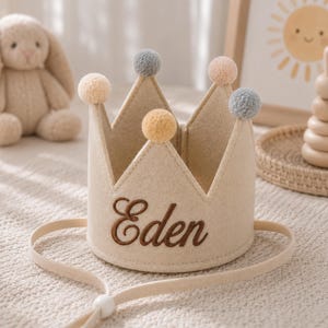 Può includere: Una corona in feltro beige con il nome "Eden" ricamato in marrone. La corona è adornata con piccoli pompon nei toni del beige e del grigio. Ha un cinturino regolabile beige. La corona è probabilmente per un bambino.