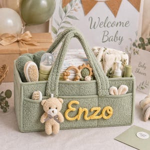 Puede incluir: Una cesta de almacenamiento de tela texturizada en color verde salvia con dos asas, que presenta el nombre Enzo en letras cursivas amarillas y un pequeño oso de peluche sujeto al frente. La cesta contiene artículos para bebé, incluyendo un biberón, cepillos, un mordedor de madera y paños suaves.