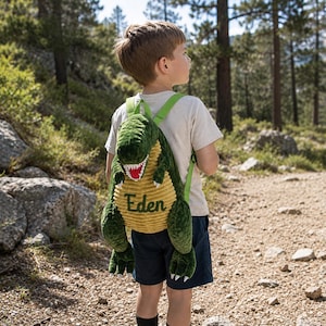 May include: A plush dinosaur-shaped backpack featuring a dark green textured exterior, a yellow corduroy belly, and a red mouth with white teeth. The name Eden is embroidered in dark green script on the belly, with adjustable light green straps.
