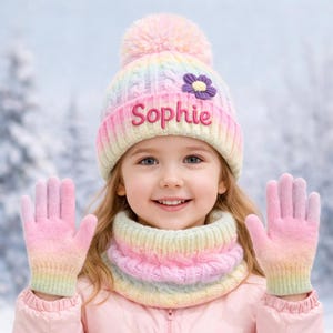 May include: A pastel-coloured winter hat, scarf, and glove set. The hat features the name "Sophie" embroidered in pink, with a purple flower. The scarf and gloves match the hat's pink, yellow, and blue colour scheme.
