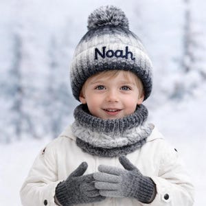 May include: A knitted winter hat, snood, and gloves set. The hat is grey and white with a dark grey pom-pom and the name "Noah" embroidered on the front. The snood and gloves are grey. The child is wearing a white jacket.